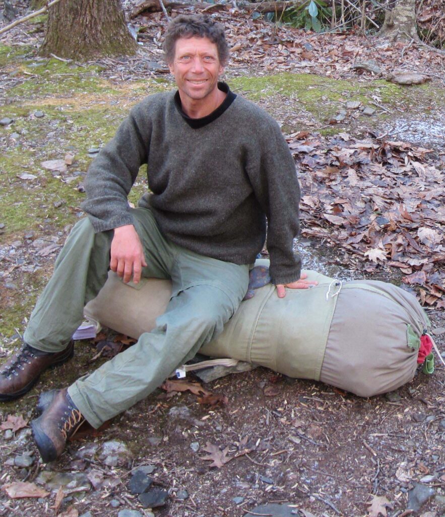 Patrick in a wool sweater sitting on a rolled wool bedroll at a winter camp in leafless woods, December 2013