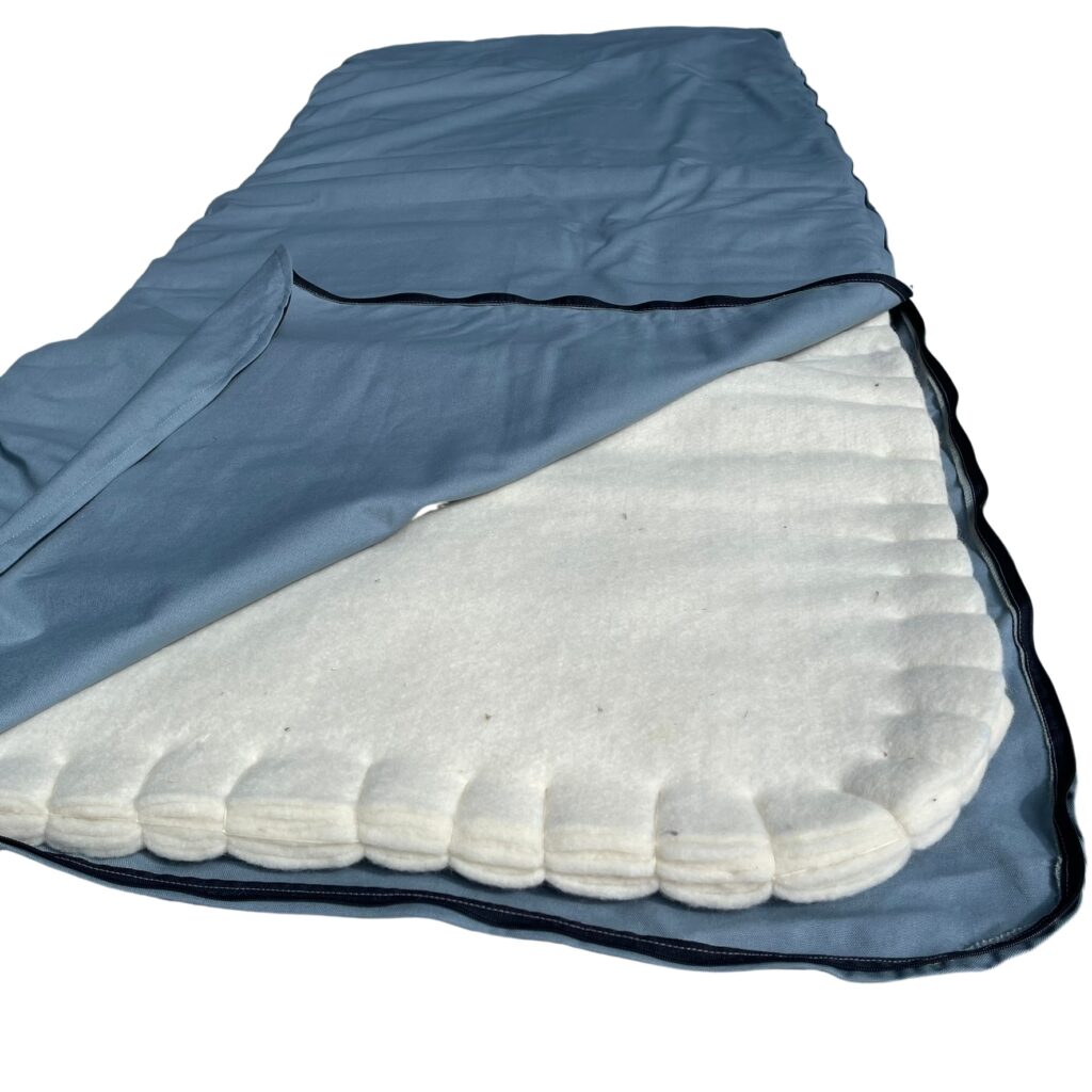 Wool sleeping pad with its blue-grey cotton cover unzipped at the foot, showing the stacked layers of cream wool felt inside