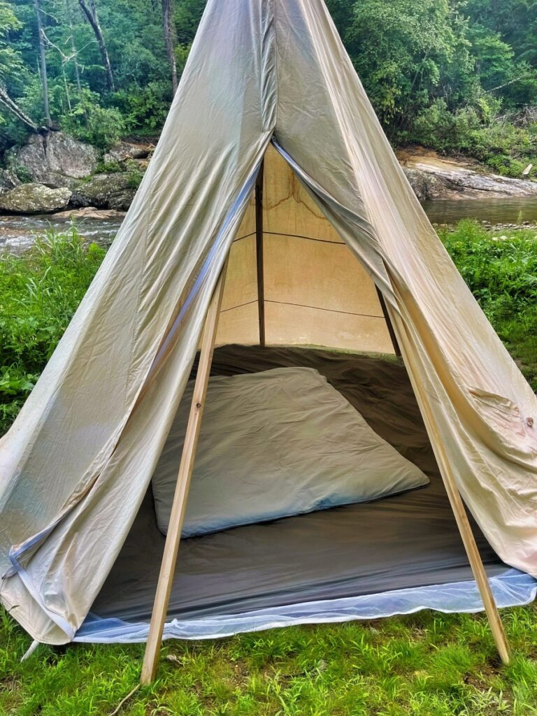 Canvas tipi tent with the door open beside a rocky creek, a grey wool sleeping pad and pillow laid out on the floor inside