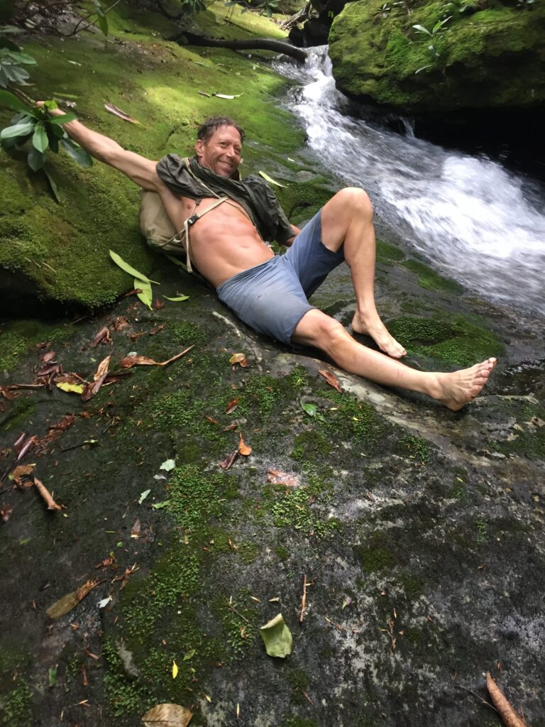 Lucky Sheep founder lying back on a moss-covered boulder beside a mountain creek, barefoot, with a small waterfall behind him