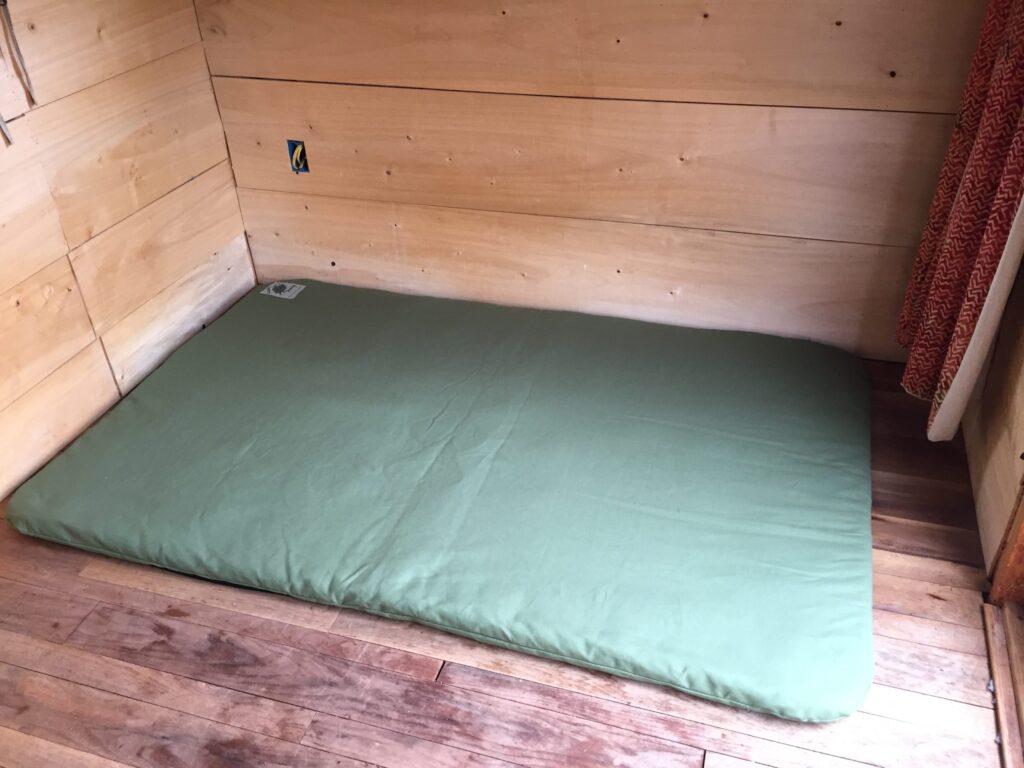 Wide green wool sleeping pad lying directly on a hardwood floor in a small pine-panelled cabin room