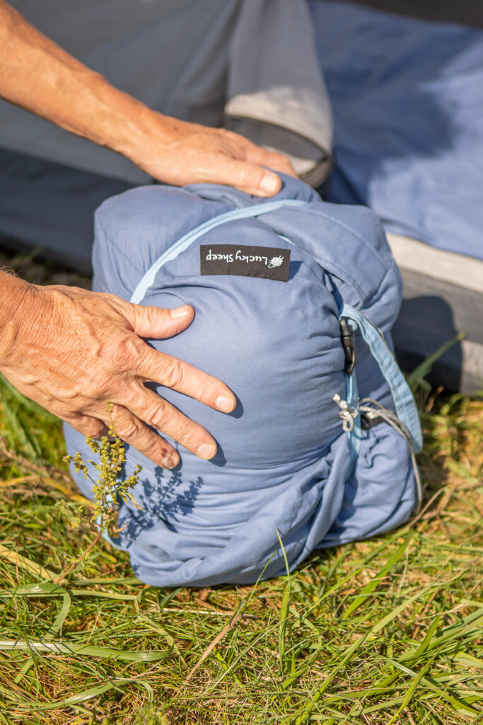 Two hands compressing a blue Rewilder wool sleeping bag with its light blue compression straps on grass beside a tent